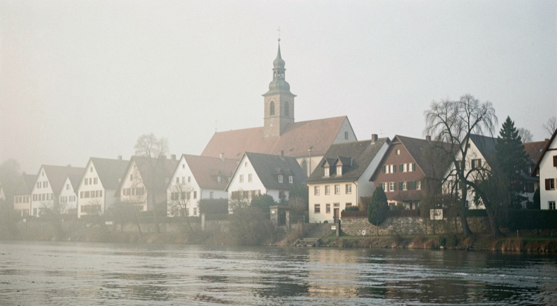 Kirchturm und Satteldächer des Dorfes von der anderen Flussseite im Nebel des frühen Morgens